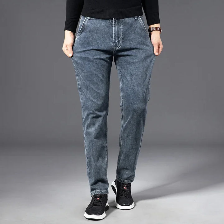 A person wearing a black sweater, straight-leg blue jeans, and black sneakers stands with hands in pockets against a plain gray background.