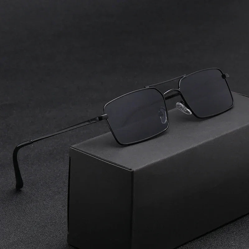 A pair of black rectangular sunglasses with thin, lightweight alloy frames rests on a black box, set against a dark, textured background.