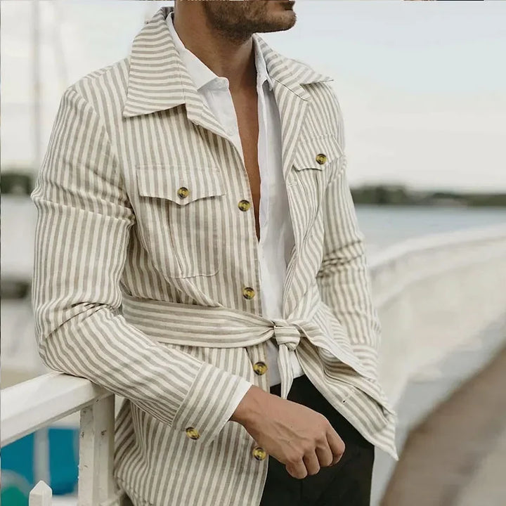 A man in a tan and white striped belted jacket with a tailored fit over a white shirt stands outdoors by a white railing, with water and trees softly blurred in the background. His face is not shown.
