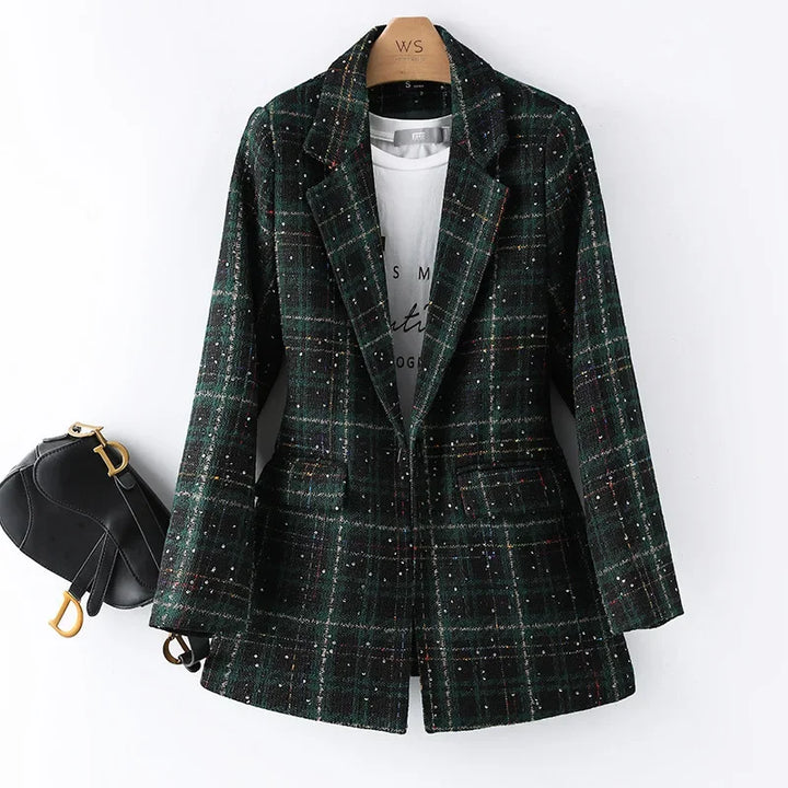 A green and black plaid formal blazer with subtle colored specks is hung on a hanger over a white printed t-shirt. A small black handbag with gold hardware is attached to the left sleeve, perfect for an office work suit look.