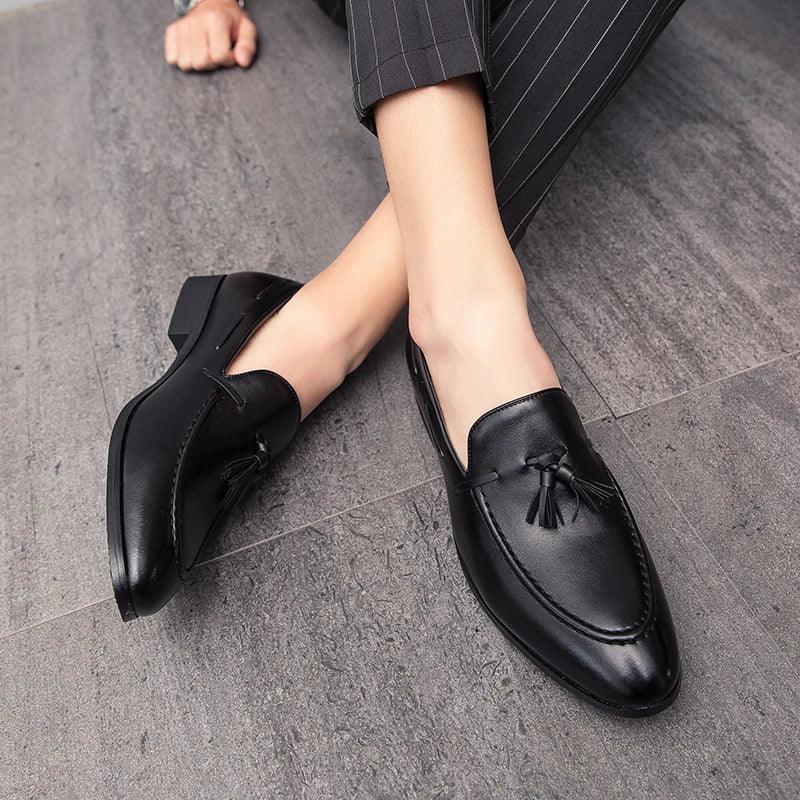 Person wearing black genuine leather mens loafers with tassels, sitting on a gray tiled floor. The person is dressed in dark pinstripe pants, and only their legs and feet are visible.