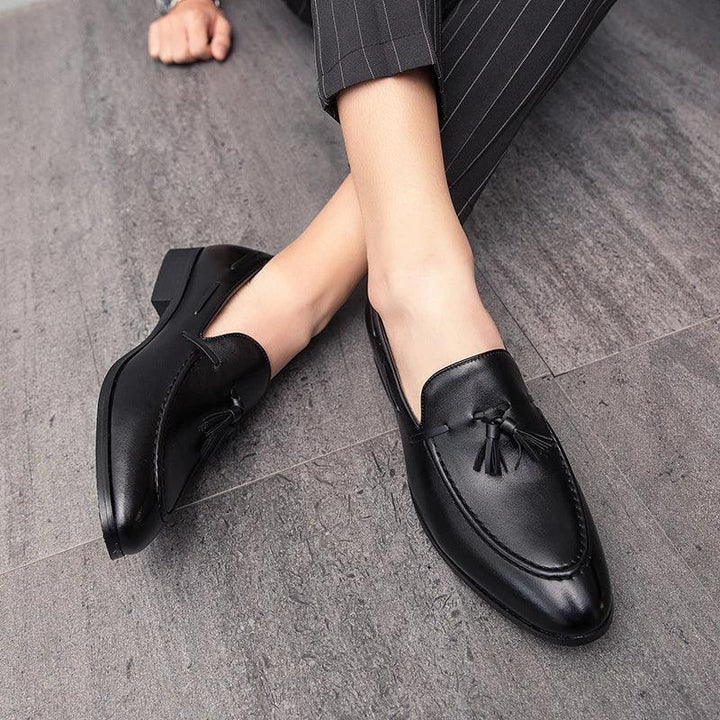 Person wearing black genuine leather mens loafers with tassels, sitting on a gray tiled floor. The person is dressed in dark pinstripe pants, and only their legs and feet are visible.