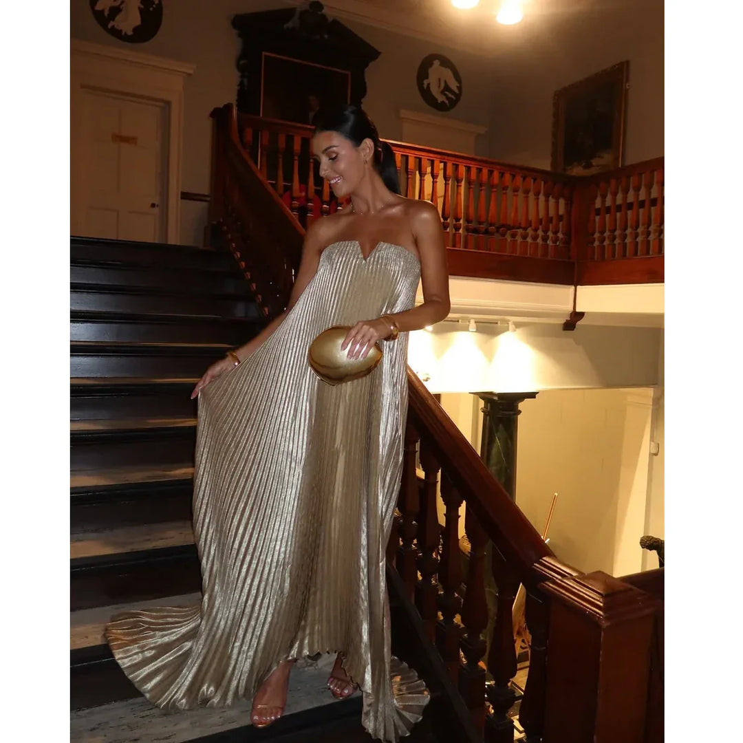 Elegant woman in gold pleated dress holding clutch on grand staircase, Rovelli evening fashion