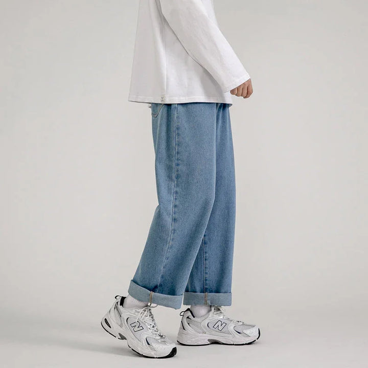 A person stands against a plain background, wearing a white long-sleeve shirt and Korean Fashion-inspired men’s baggy jeans with rolled cuffs, paired with white socks and New Balance sneakers.