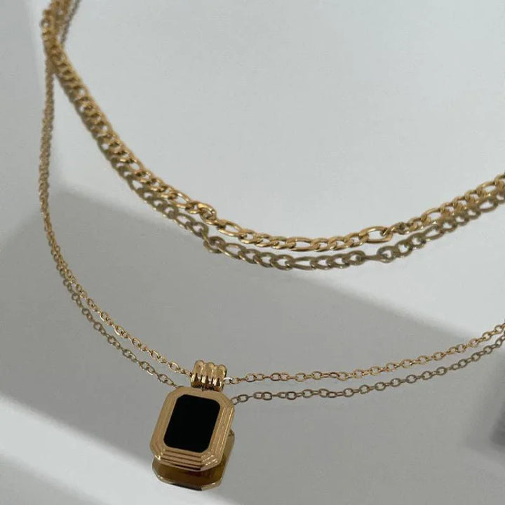 Two gold chain necklaces on a white surface; one necklace has a simple stainless steel chain, while the other features a rectangular black pendant set in gold. Shadows of the chains are visible in the background.