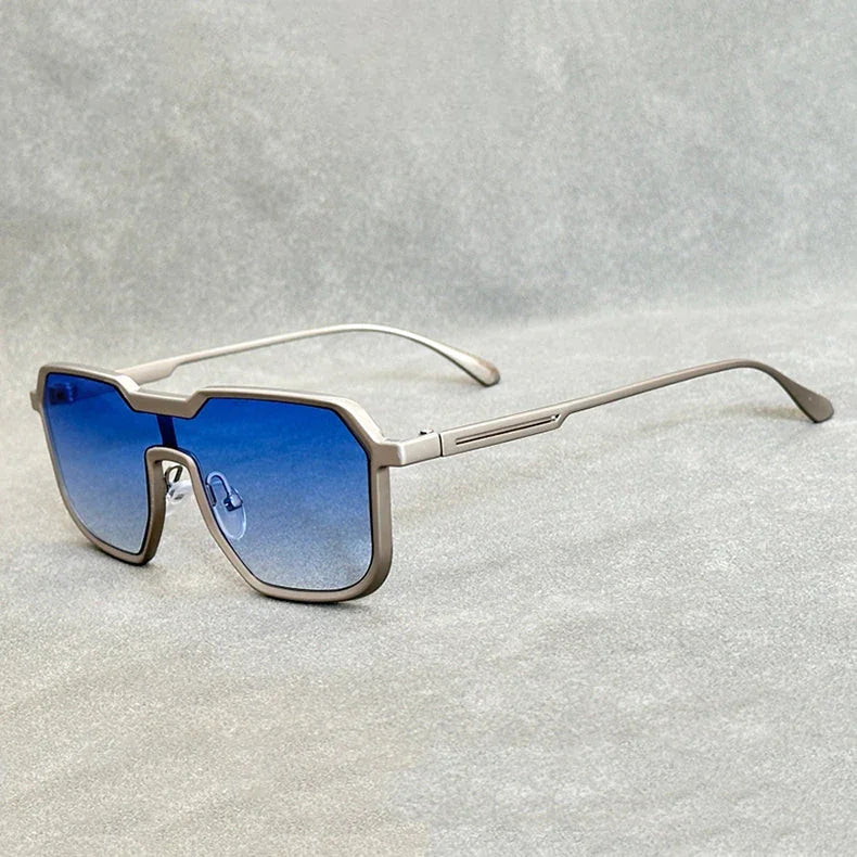 A pair of modern, geometric Heavy Metal Sunglasses with square metallic frames and blue gradient lenses rests on a light grey textured surface.