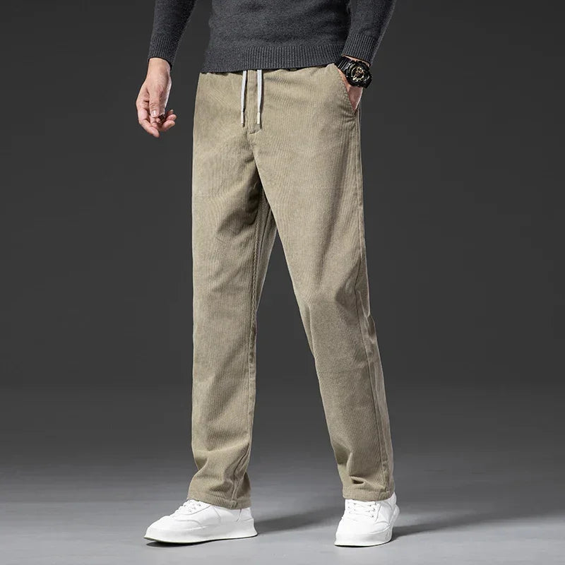 A person wearing men's smart casual corduroy pants with a zipper fly, a dark grey sweater, a watch, and white sneakers stands against a plain dark background. The photo is cropped from the shoulders down.