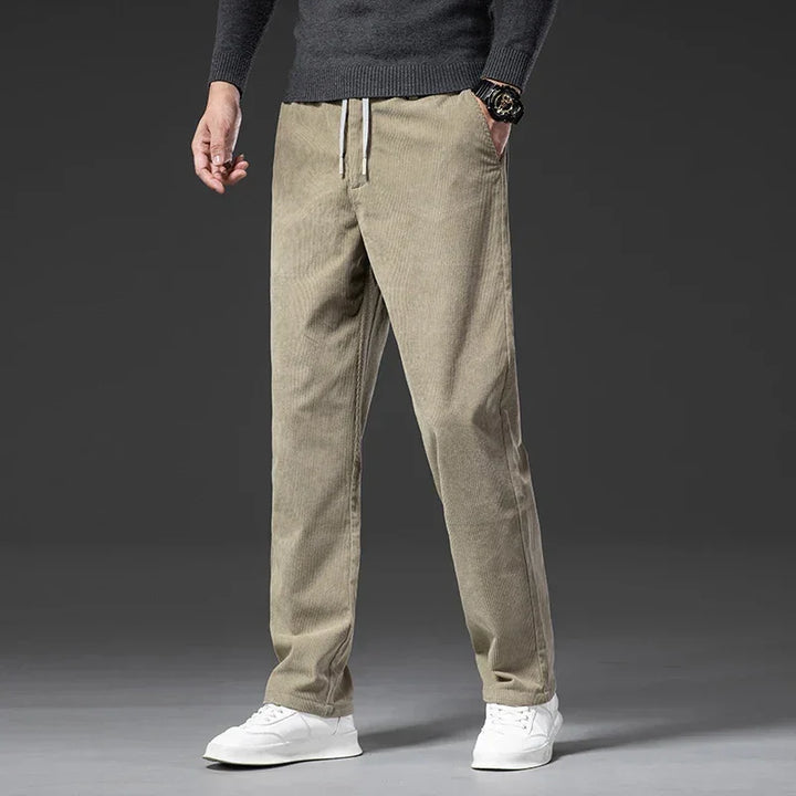 A person wearing men's smart casual corduroy pants with a zipper fly, a dark grey sweater, a watch, and white sneakers stands against a plain dark background. The photo is cropped from the shoulders down.