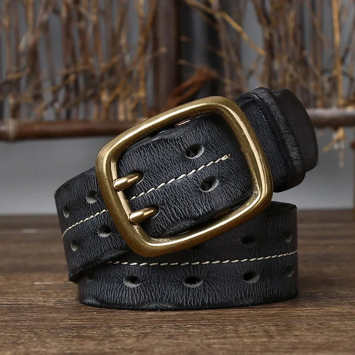 A vintage-inspired belt, this coiled black leather accessory features a gold-tone double prong buckle, white stitching along the edges, and multiple oval holes, displayed on a wooden surface with a rustic background.