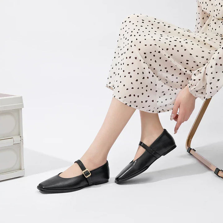 A person wearing a cream-colored, long-sleeve dress with black polka dots sits on a chair, showing stylish Mary Jane shoes with a buckled strap. Only the lower half of their body is visible.