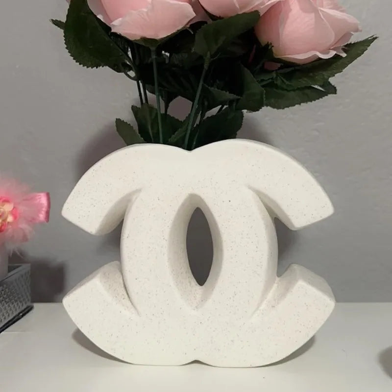 A decorative white sculpture in an Irregular INS Style, resembling the Chanel double C logo, is placed on a surface with pink artificial roses and green leaves in the background—perfect for Nordic home decoration.