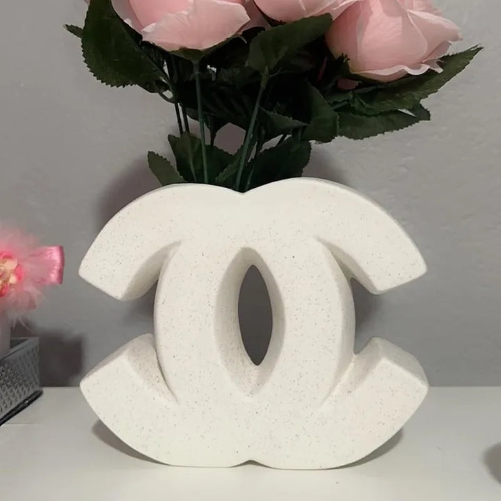 A decorative white sculpture in an Irregular INS Style, resembling the Chanel double C logo, is placed on a surface with pink artificial roses and green leaves in the background—perfect for Nordic home decoration.