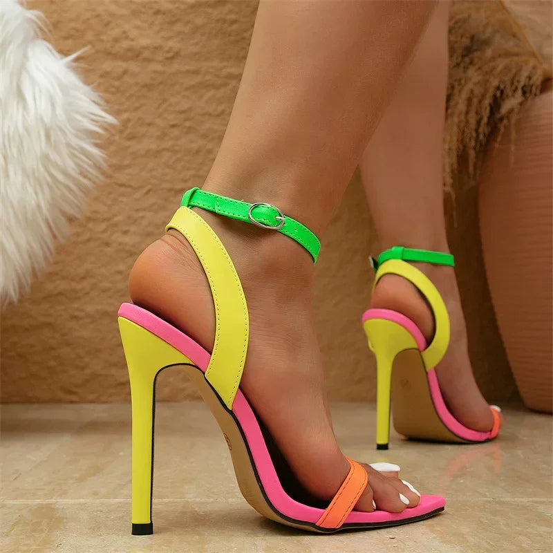 A person wearing high-heeled sandals with a fashion-forward design featuring neon yellow, green, pink, and orange straps and an adjustable ankle strap; their toenails are painted white as they stand indoors on a marble floor.