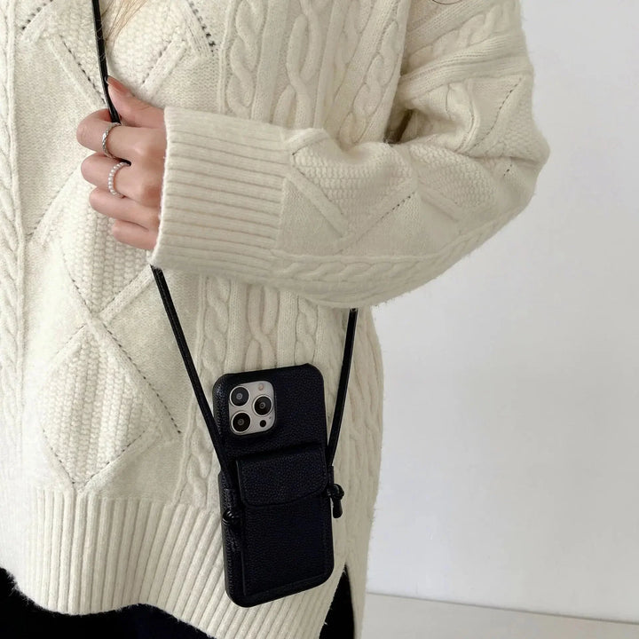 A person wearing a cream-colored cable knit sweater holds a black leather phone case with a detachable lanyard. The case, hanging from their neck, contains a smartphone with three cameras.