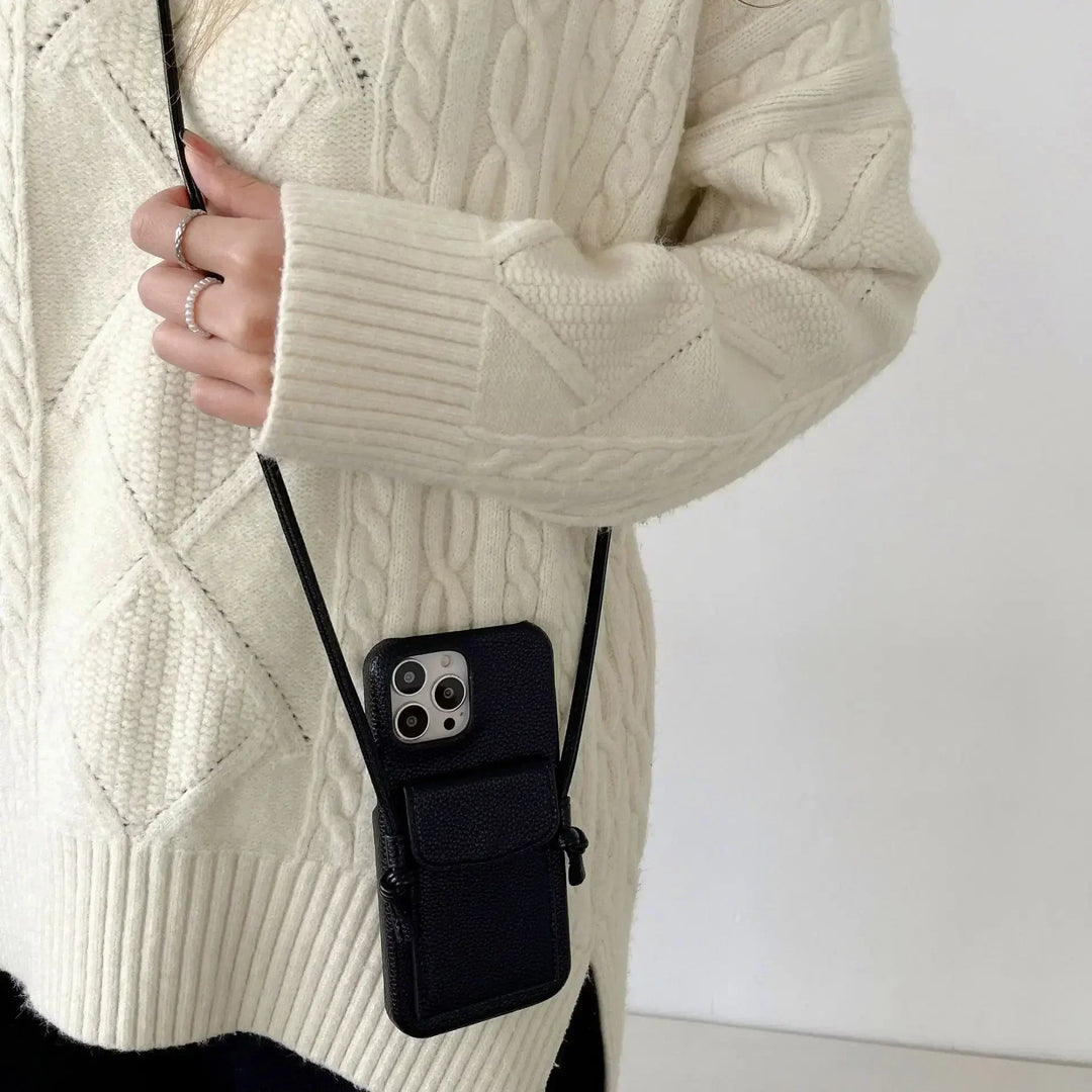 A person wearing a cream-colored cable knit sweater holds a black leather phone case with a detachable lanyard around their neck; only their torso and hand are visible.