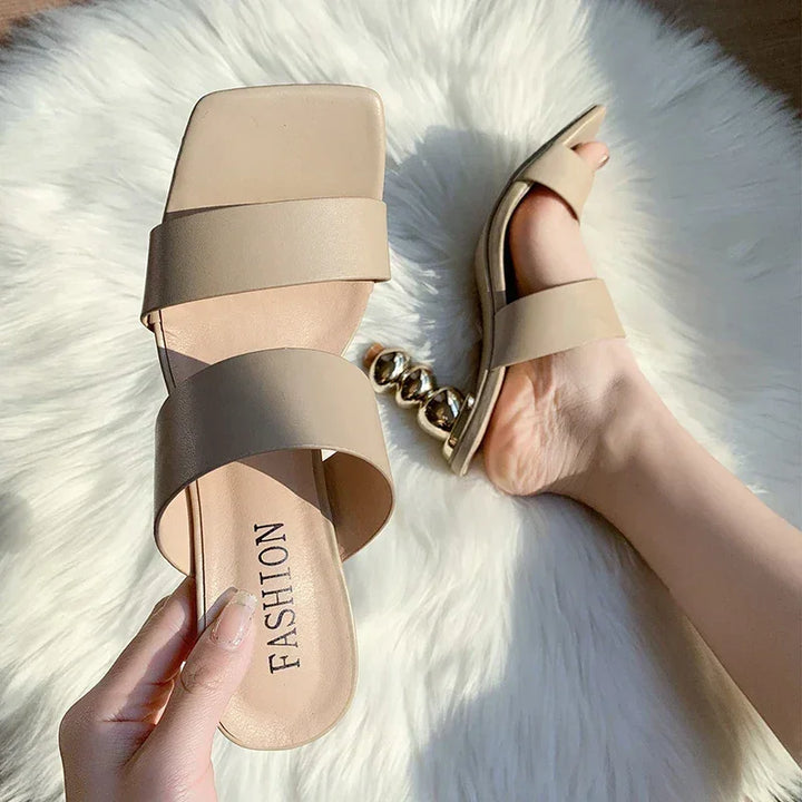 A person holds a beige sandal with two wide straps and a square toe featuring a cushioned footbed; another sandal with metallic ball-shaped sculpted heel is worn on their foot, both resting on a fluffy white rug.