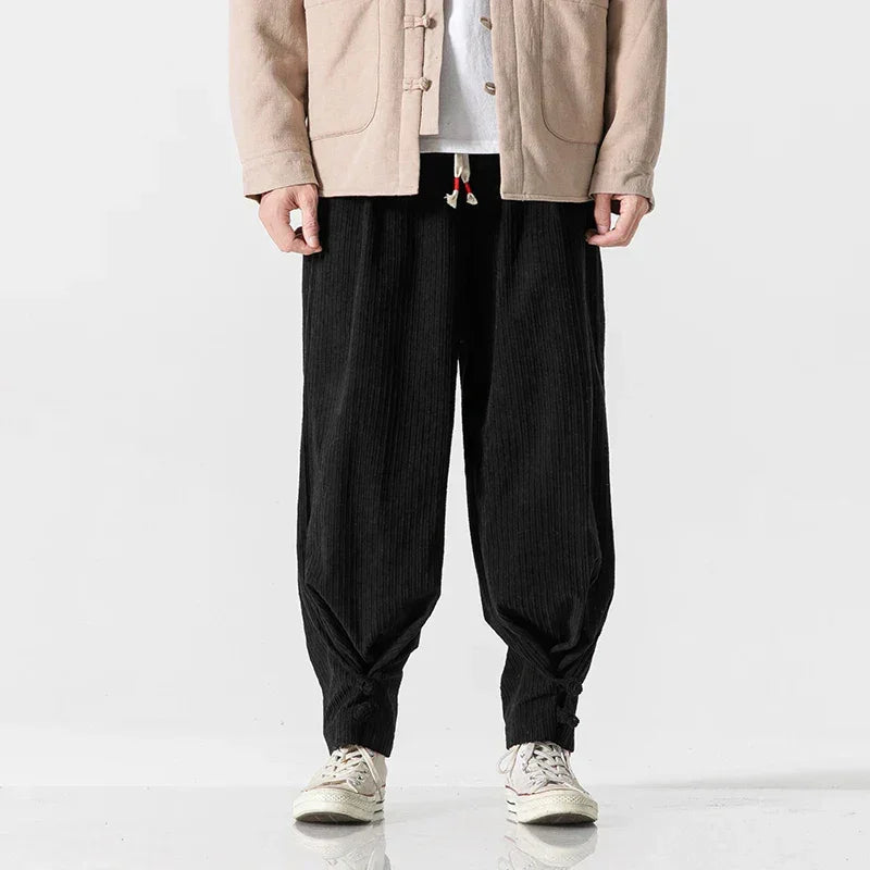 A person wears a beige jacket, white shirt, and black cotton blend sweatpants featuring a drawstring waistband and adjustable fit at the ankles, paired with white sneakers, standing against a plain white background. The persons head is not visible.