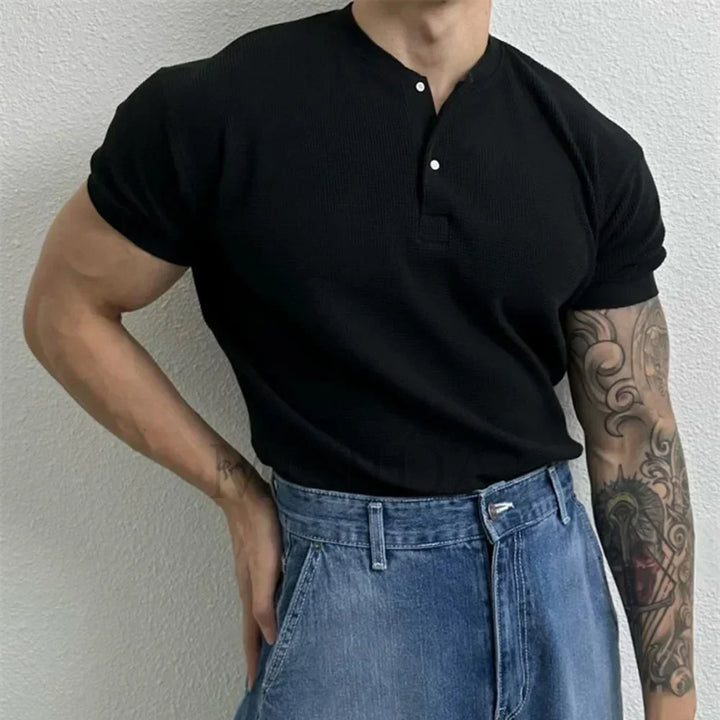 A person stands against a light-colored wall, wearing a black ribbed Henley tucked into high-waisted blue jeans. One arm is bent at the waist, revealing a detailed tattoo sleeve.