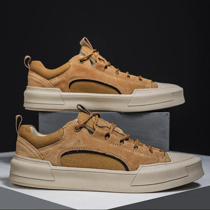 A pair of tan and brown suede canvas shoes with thick white soles are displayed, one shoe elevated on a gray block and the other resting on a flat surface, against a dark background.