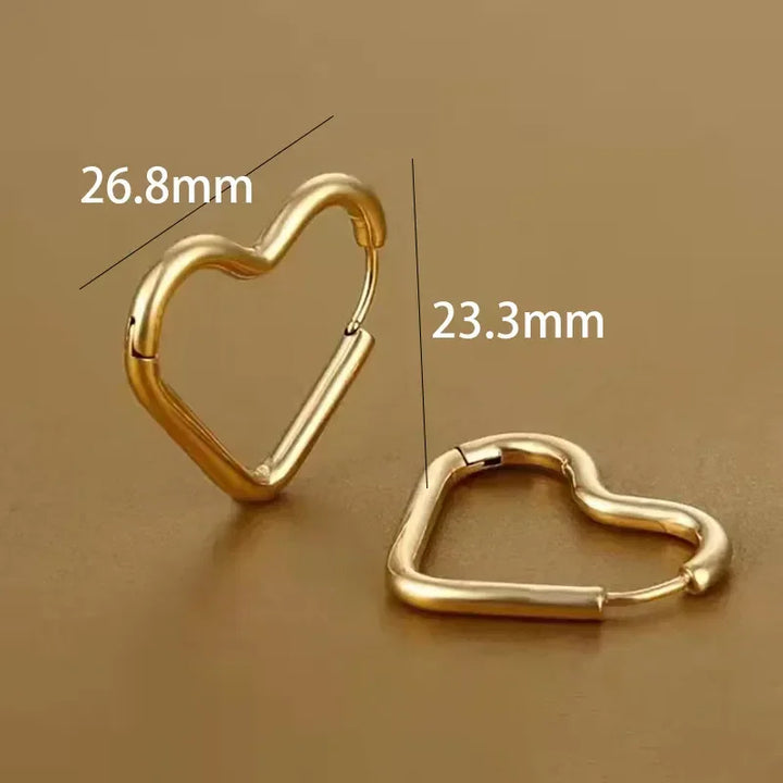 Two gold heart-shaped Eternal Love Earrings rest on a brown surface; one stands upright at 26.8 mm high, the other lays flat showing a 23.3 mm width—highlighting their elegant craftsmanship and minimalist design.