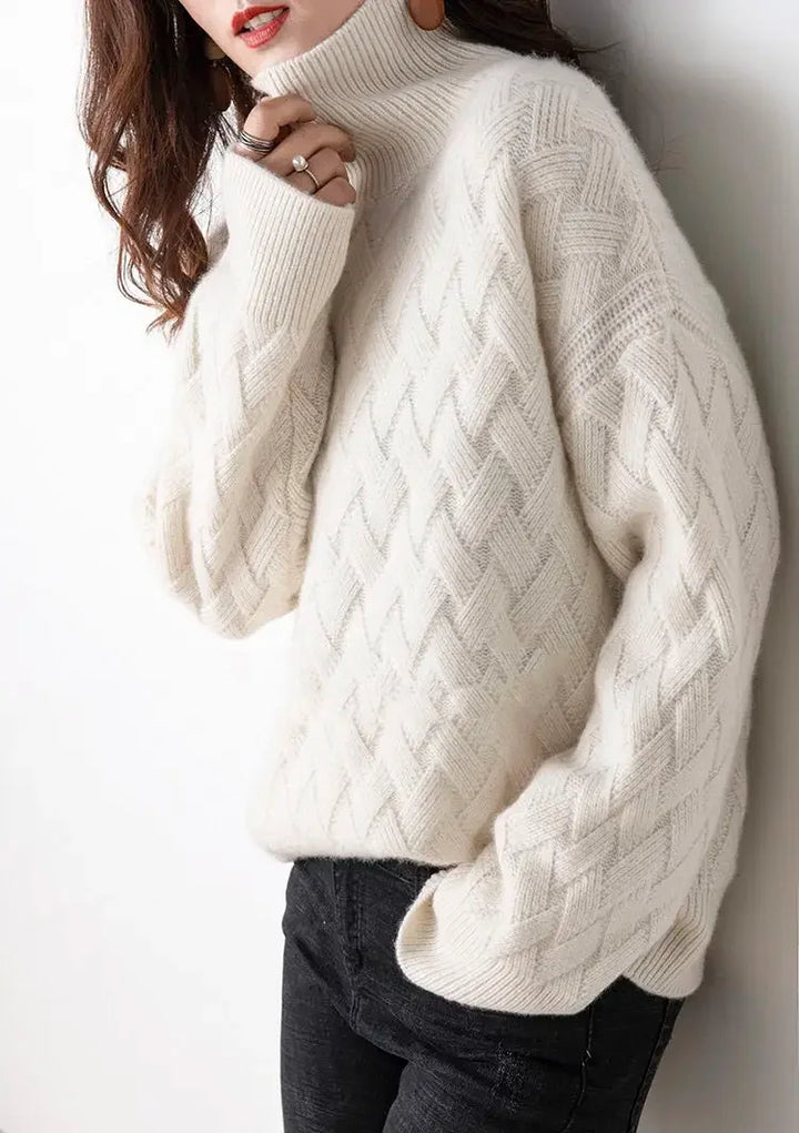 A woman wearing an ultra soft white cable-knit turtleneck sweater and black pants stands against a white background, with one hand touching her chin and her long hair falling over her shoulder.