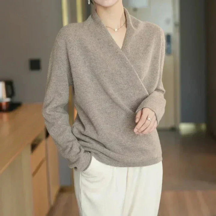 A person wearing a light brown, cashmere wrap-style sweater and cream-colored pants stands indoors with one hand in their pocket. Their head is partly out of frame.