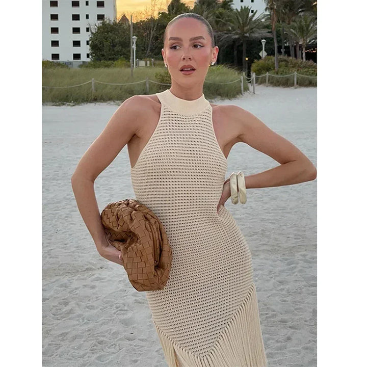 Woman on beach wearing sleeveless crochet beige dress, holding woven bag, sunset background Rovelli