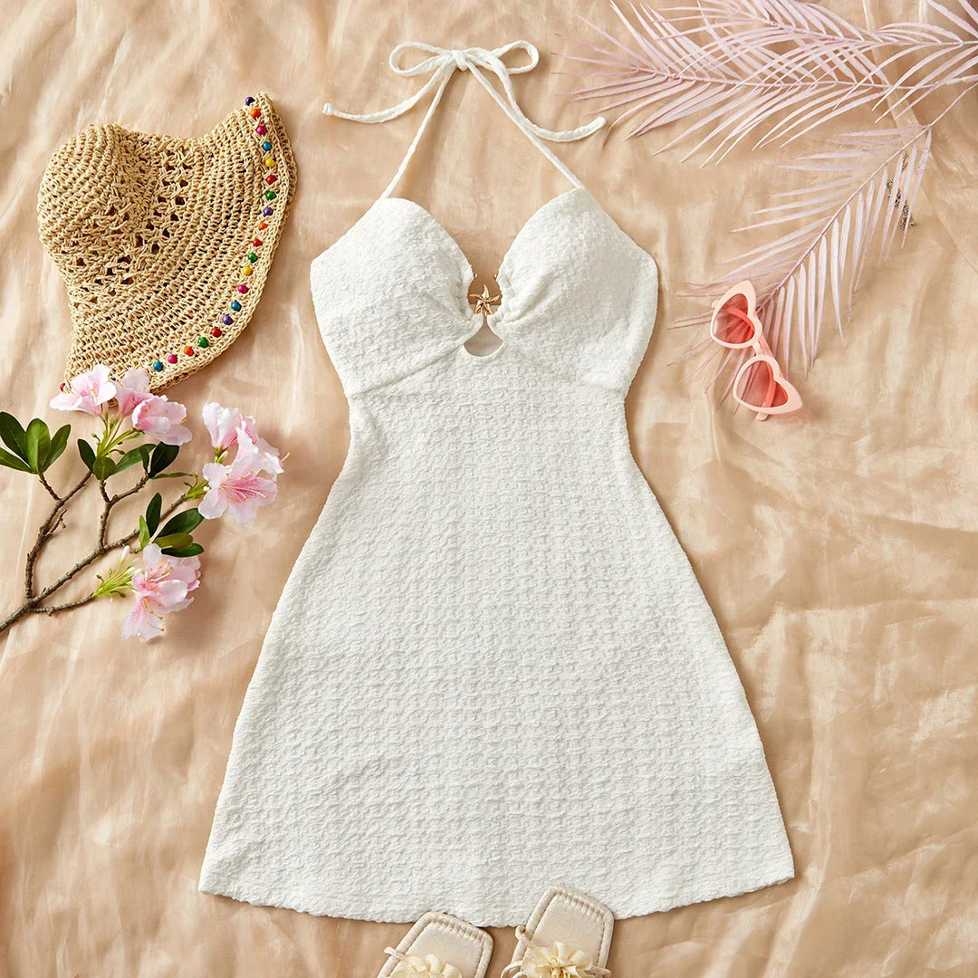 White halter dress flat lay with straw hat, pink heart sunglasses, and floral accents from Rovelli
