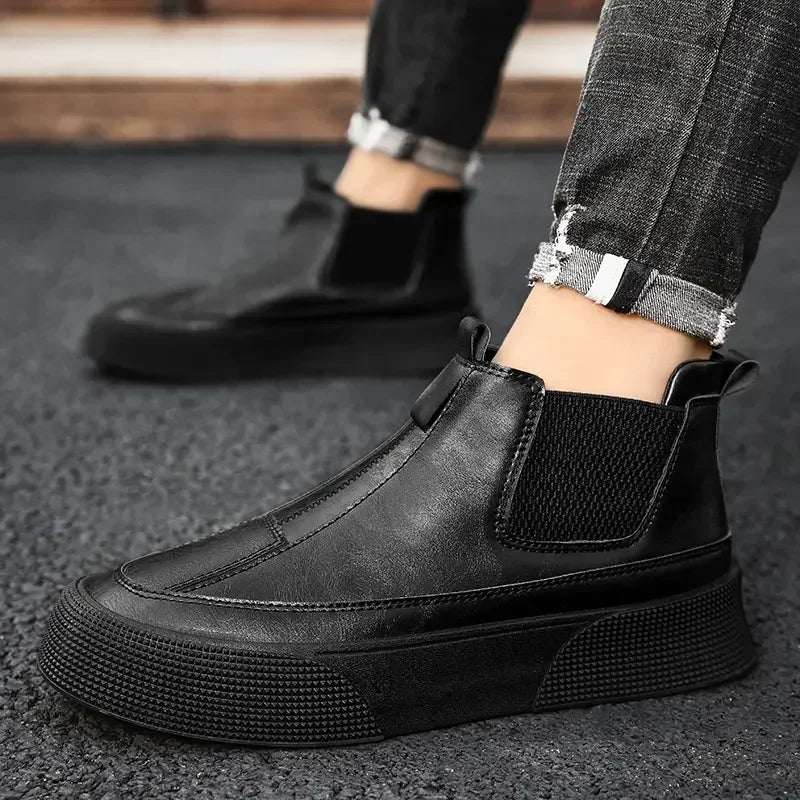 A person wearing black slip-on sneakers with a cushioned footbed and thick soles stands on a textured dark surface, dressed in cuffed, distressed black jeans.