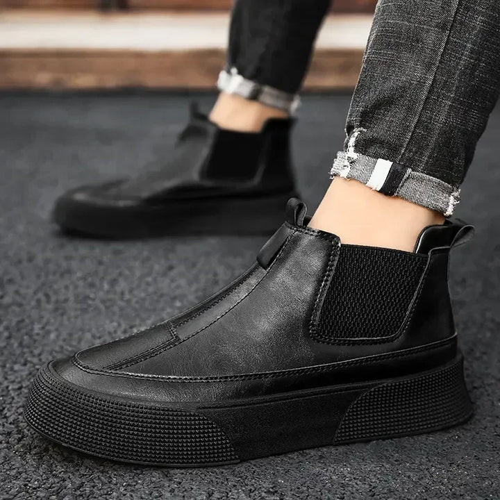 A person wearing black slip-on sneakers with a cushioned footbed and thick soles stands on a textured dark surface, dressed in cuffed, distressed black jeans.