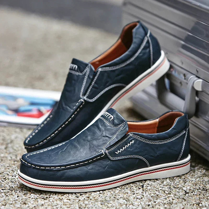 A pair of navy blue boat-style leather loafers with white stitching and white-red rubber soles sits on a gravel surface next to a silver suitcase.