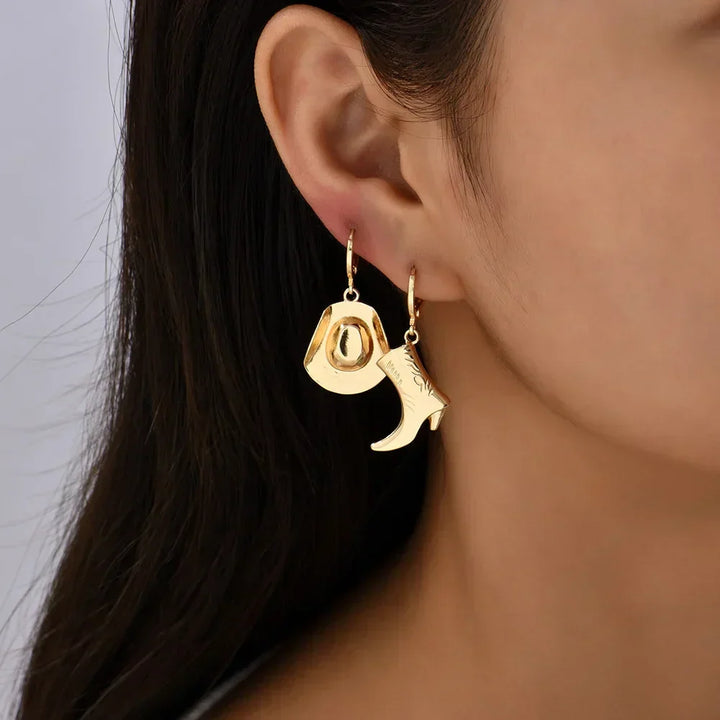 Close-up of a woman’s ear adorned with mismatched gold earrings—a cowboy hat and a boot—showcasing stylish western jewelry, with long dark hair partially visible.