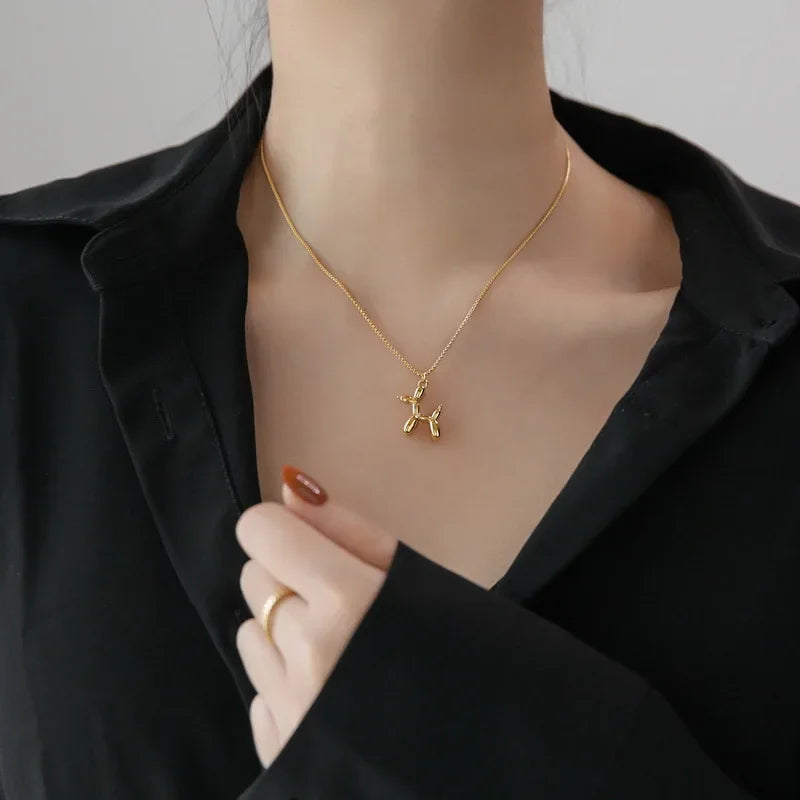 A person wearing a black shirt displays a balloon dog necklace with a gold pendant. Their hand, adorned with a gold ring and brown nail polish, gently touches the shirt near the collar, highlighting this playful jewelry piece.