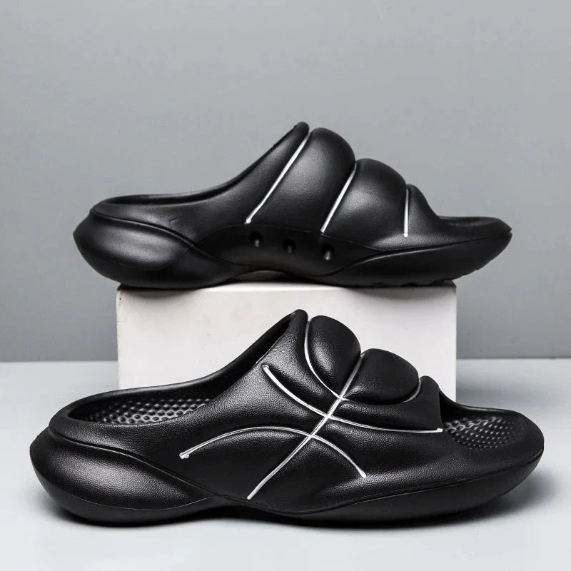 A pair of black basketball slip ons with thick, cushioned soles and white curved line designs are displayed, one on a white box and the other on the surface, against a plain gray background.
