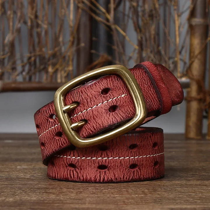 A brown textured genuine leather belt with oval-shaped holes and a brass buckle is coiled on a wooden surface, set against a rustic background of dried branches and bamboo sticks, highlighting heritage craftsmanship.