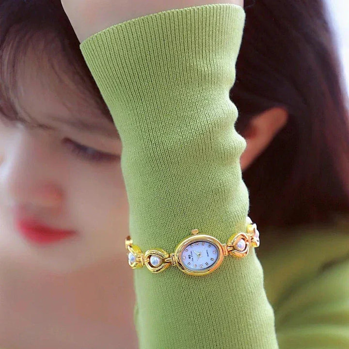 A woman in a light green long-sleeve top covers her face with her arm, showcasing a gold and pearl accents bracelet watch on her wrist. Her softly blurred features create an elegant backdrop for the timepiece.