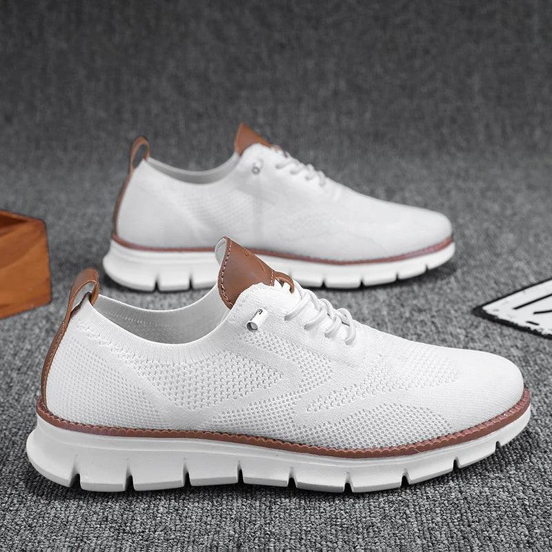 A pair of white knit, breathable loafers with brown accents on the tongue and heel, featuring thick, white, cushioned soles. The shoes are displayed on a gray textured surface.