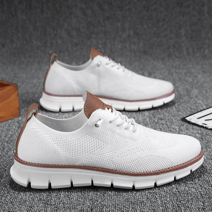 A pair of white knit, breathable loafers with brown accents on the tongue and heel, featuring thick, white, cushioned soles. The shoes are displayed on a gray textured surface.