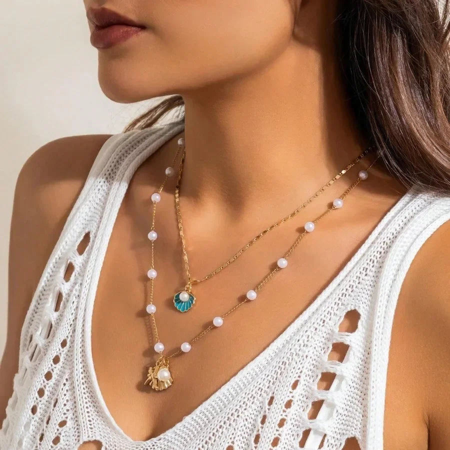 Woman wearing layered gold necklaces with pearl and turquoise shell pendants, white crochet top, Rovelli