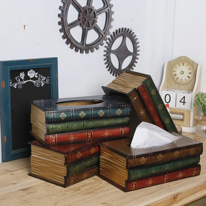 A stack of decorative tissue boxes designed to look like vintage book tissue boxes sits on a wooden desk, with gears, a small chalkboard, and a clock in the background. One tissue partially emerges from a book-shaped box.