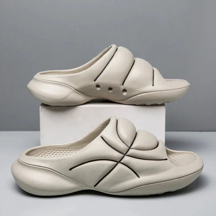 Two beige slide sandals with a cushioned footbed and chunky design feature black curved line accents, one resting on a white box and the other on the table, against a plain gray background.