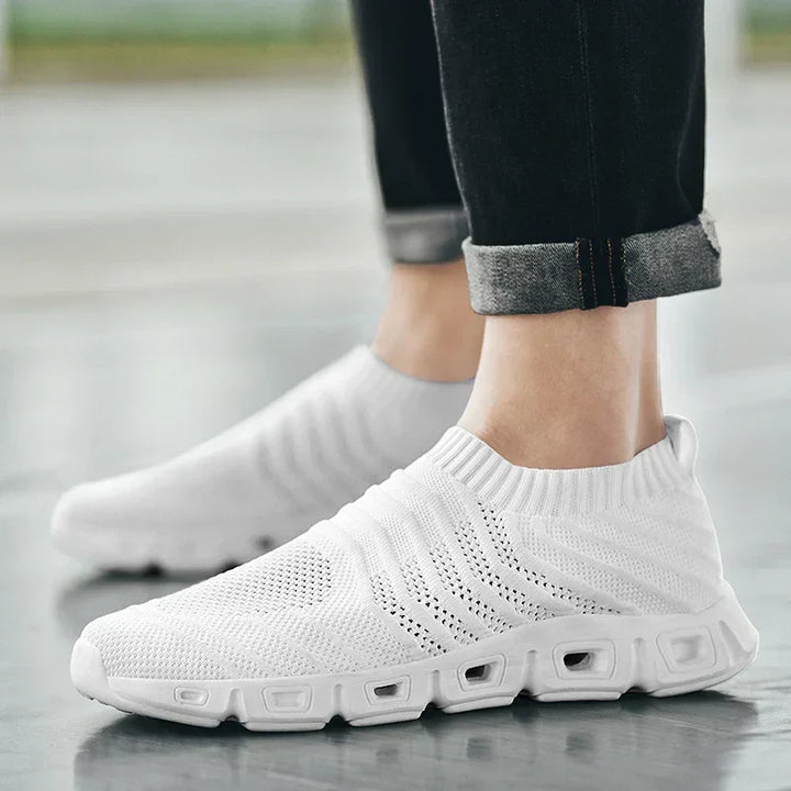 Person wearing white slip-on athletic sneakers with a textured knit upper, cushioned sole, and chunky design, standing indoors on a smooth surface. Pants are black and rolled up at the cuffs.