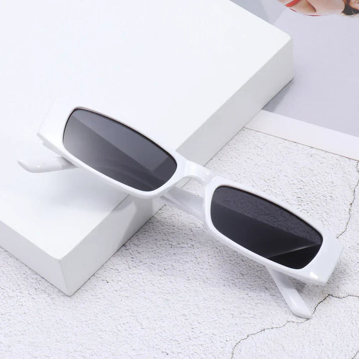 A pair of retro rectangle sunglasses with a lightweight frame and dark UV400 protection lenses rests on a light textured surface near a white box and a partially visible magazine.
