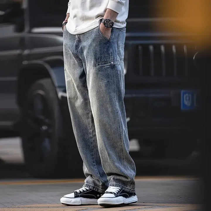 A person wearing loose-fitting, classic denim blue jeans, a white long-sleeve shirt, a watch, and black-and-white sneakers stands outdoors with hands in pockets, in front of a dark-colored vehicle.