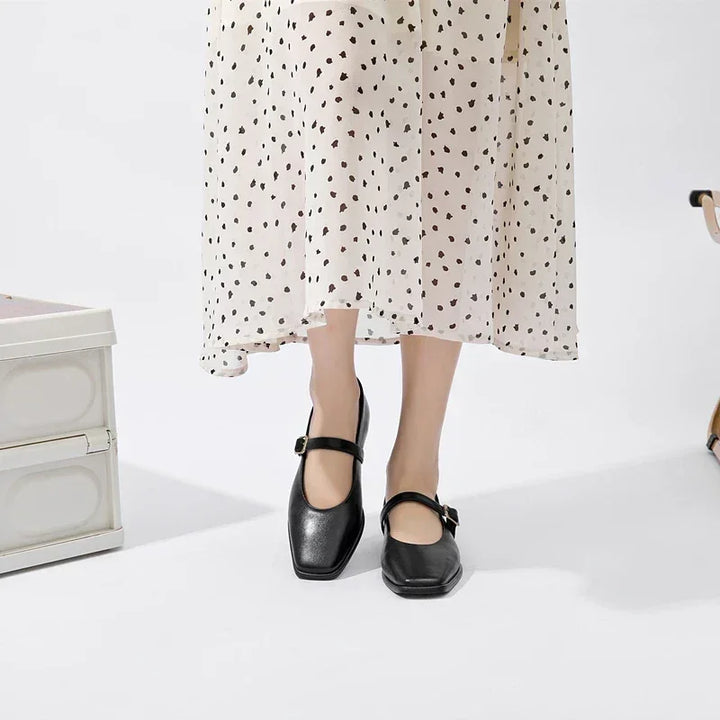 A person wearing a cream skirt with black polka dots and black ballet flats stands on a white floor. Part of a white storage box and a chair are visible nearby.