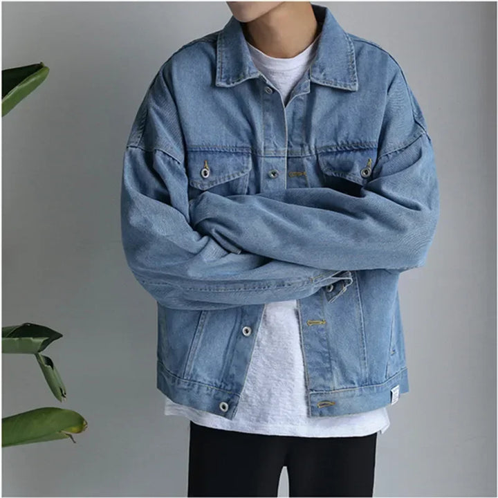 Person standing with arms crossed, wearing an oversized light blue premium denim jacket over a white T-shirt and black pants, with a neutral gray background and a plant partially visible on the left side.