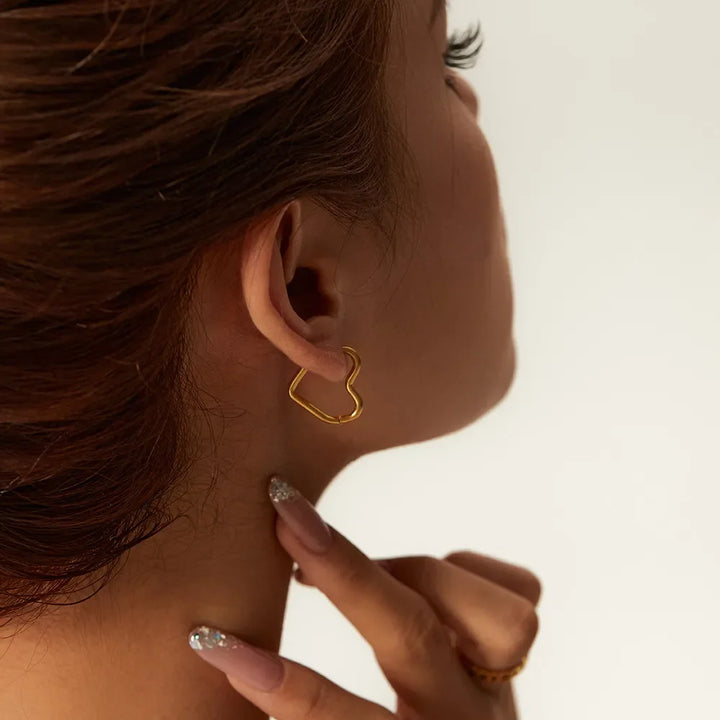 A person with brown hair, seen from the side, wears an Eternal Love Earring with a minimalist design, touching their neck with manicured fingers. The background is plain and light-colored.