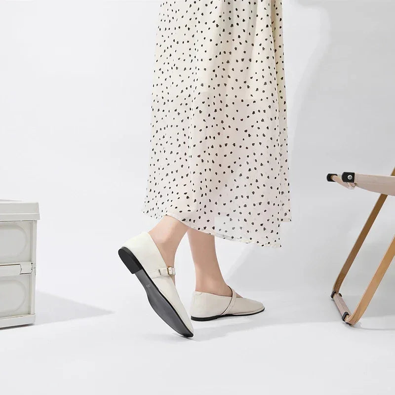 A person wearing a white dress with black dots and cream-colored ballet flats stands next to a beige folding chair and a light-colored storage box on a white background.