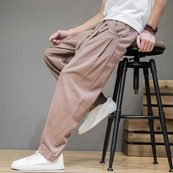 Man wearing loose brown pleated pants and white sneakers sitting on a stool, Rovelli fashion