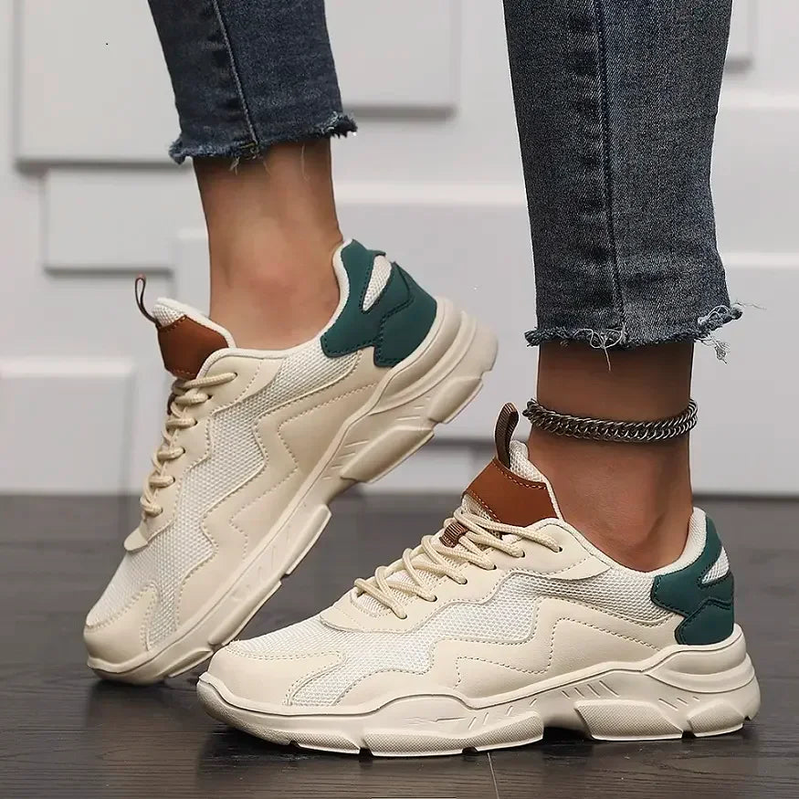A person wearing beige chunky slip-on street shoes with green accents and brown details, featuring advanced shock absorption, paired with frayed-edge skinny jeans and a silver anklet, standing on a dark wooden floor against a white textured wall.